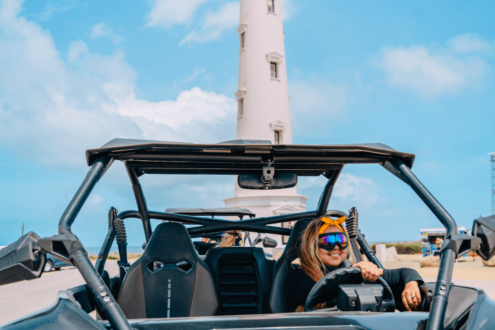 Person driving an off-road vehicle in front of a tall white lighthouse under a blue sky.