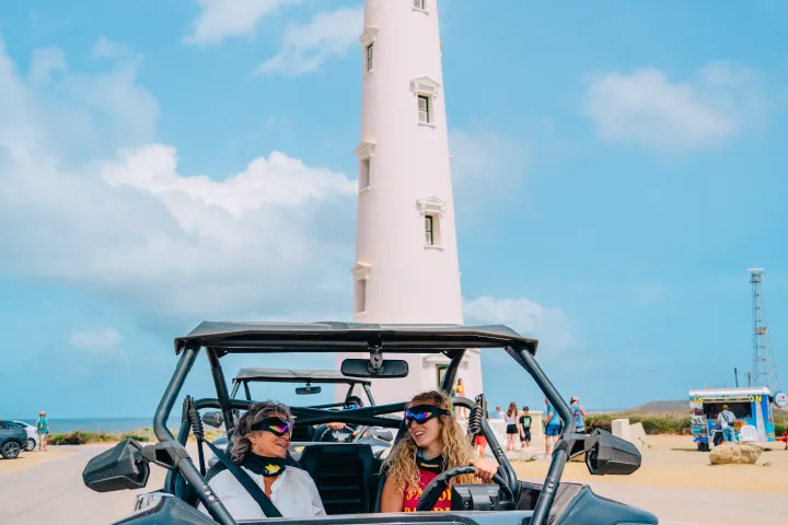 Two people in a dune buggy near a tall lighthouse on a sunny day.
