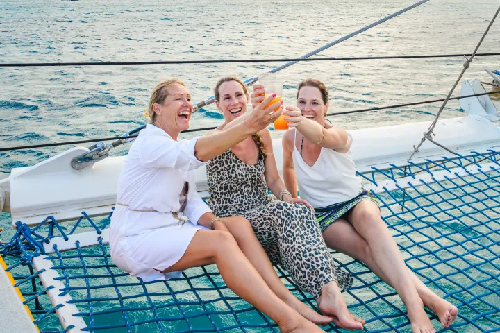 Three women toasting drinks on a boat's netting over the water.