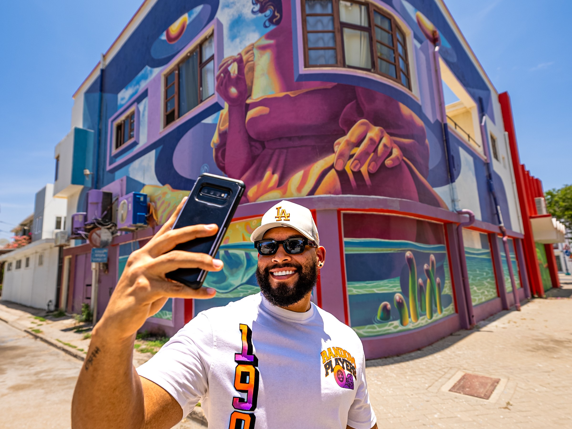 Man taking a selfie in front of a colorful mural on a sunny day.