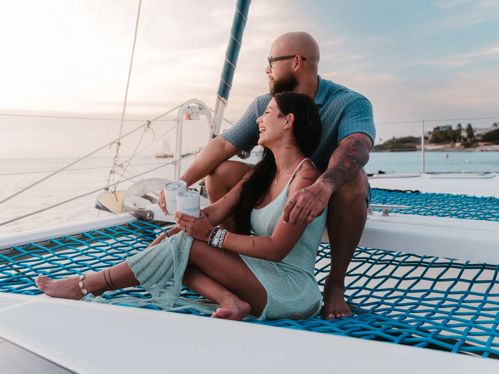 Couple sitting on a catamaran netting, smiling and holding a drink, with a sunset over the ocean.