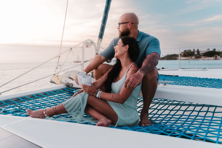 Couple sitting on a catamaran netting, smiling and holding a drink, with a sunset over the ocean.