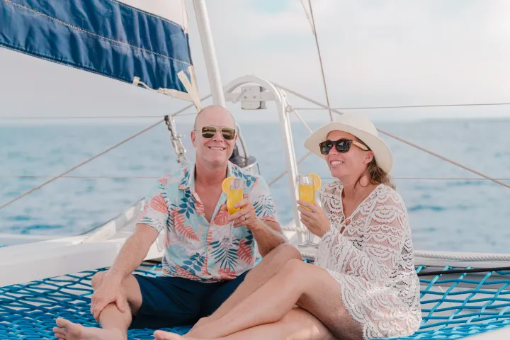 Couple on sailboat net, wearing sunglasses and holding drinks, under blue sky.