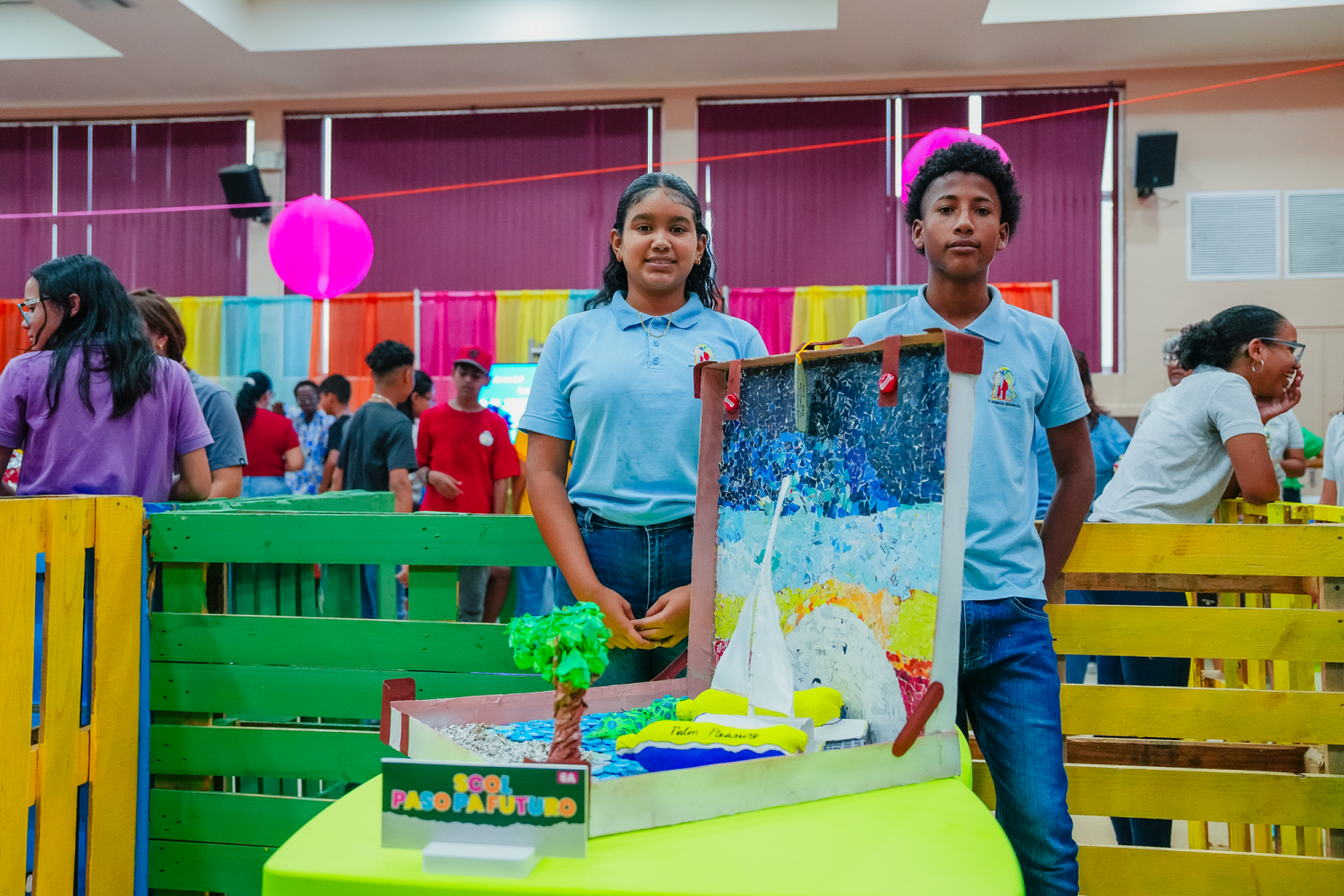Two students in blue shirts stand by a colorful school project in a decorated room.