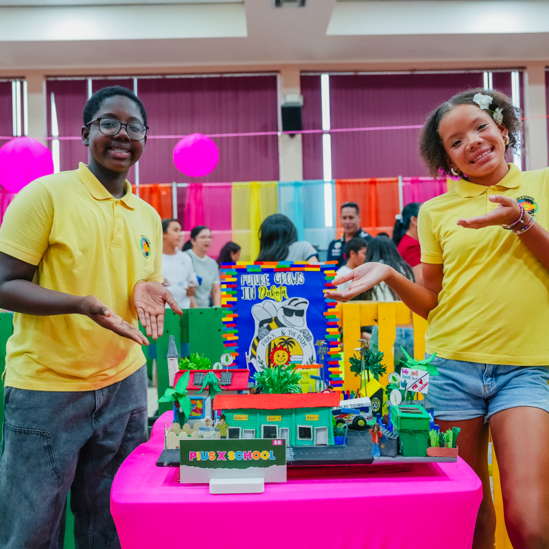 Two children in yellow shirts presenting a colorful miniature diorama at an indoor event.