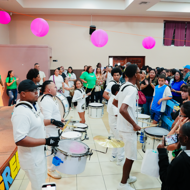 Drumline in white uniforms performing for an audience in a decorated hall with pink lanterns.