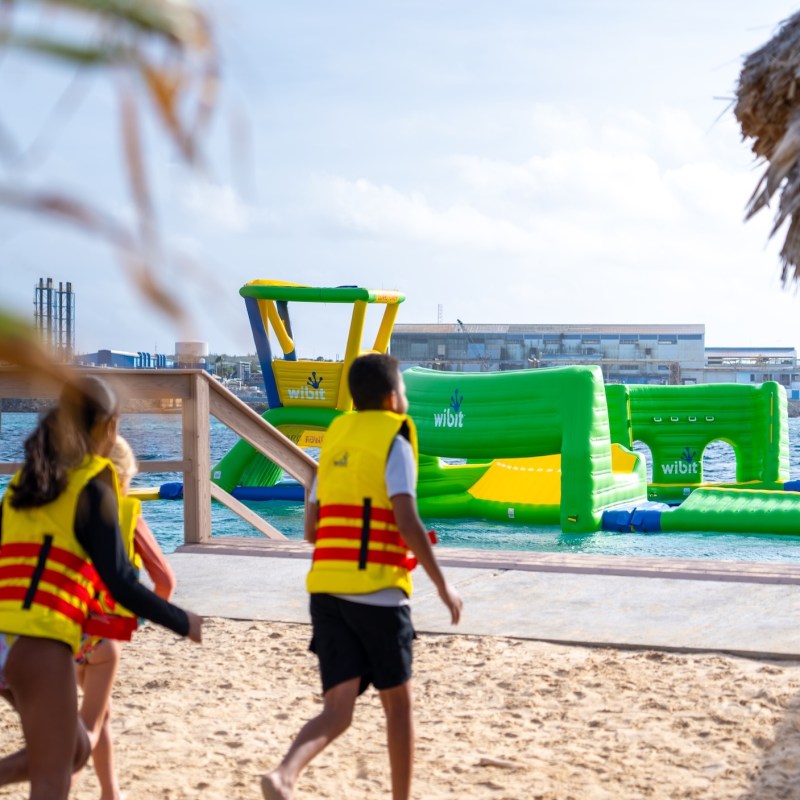 Children in yellow life jackets running toward an inflatable water park on a sunny beach.