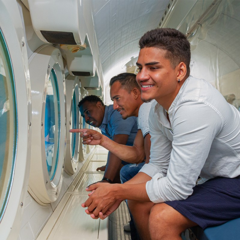 Three men inside a submarine looking out through portholes, smiling and pointing.