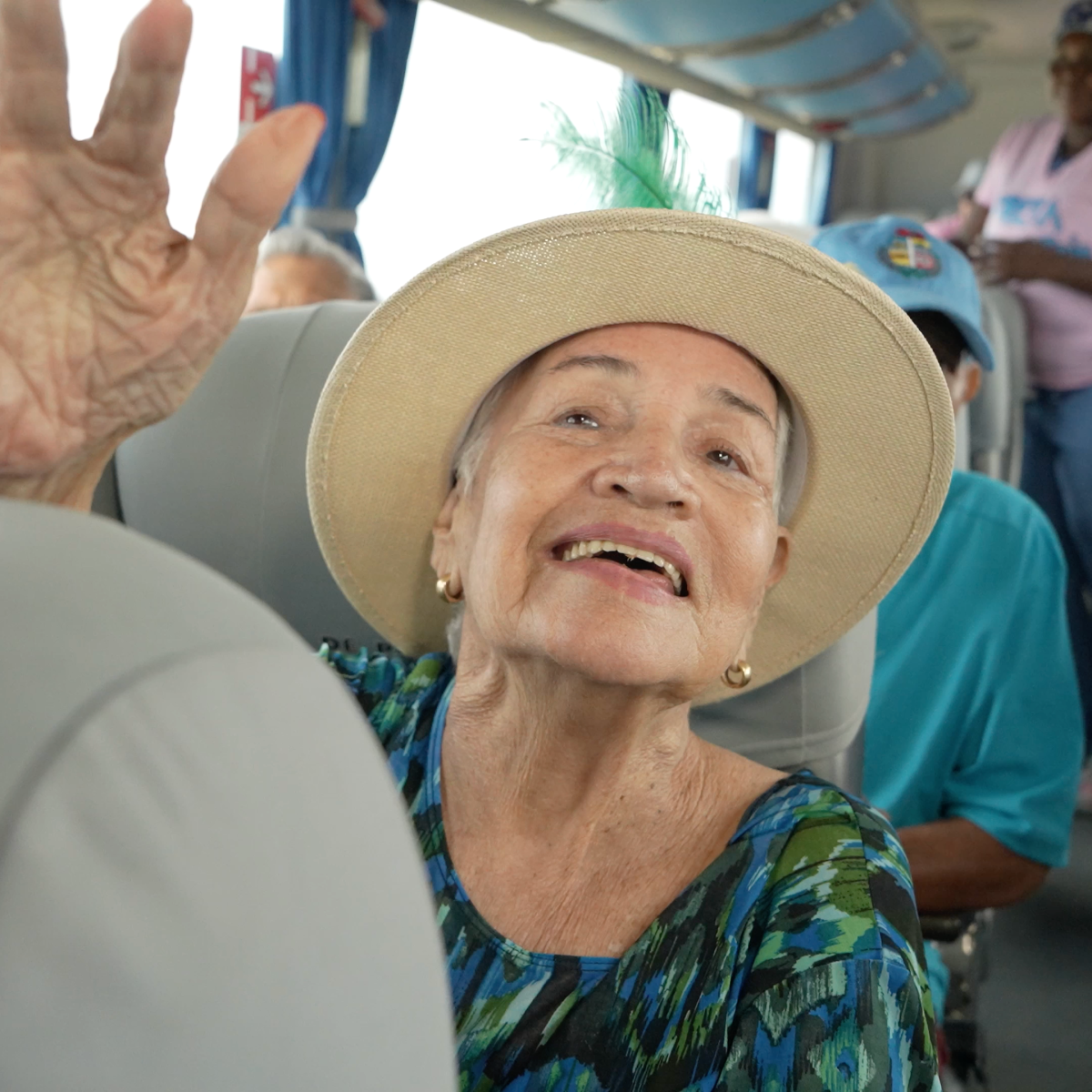 Smiling elderly woman in a hat waves inside a bus with other passengers.