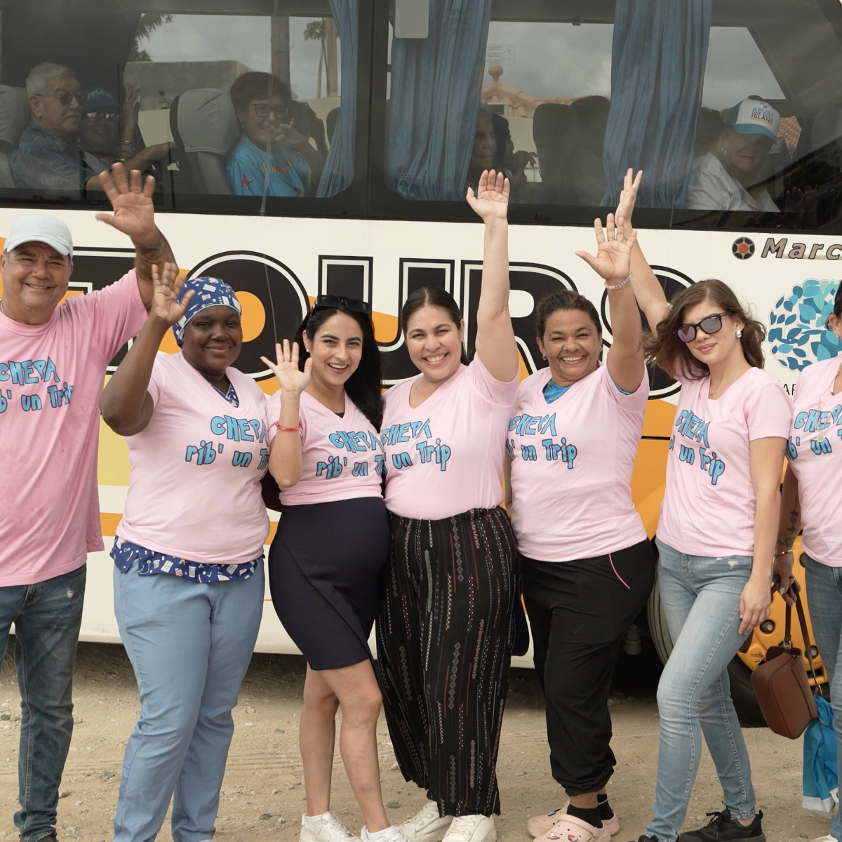 Seven people in pink shirts wave in front of a tour bus.