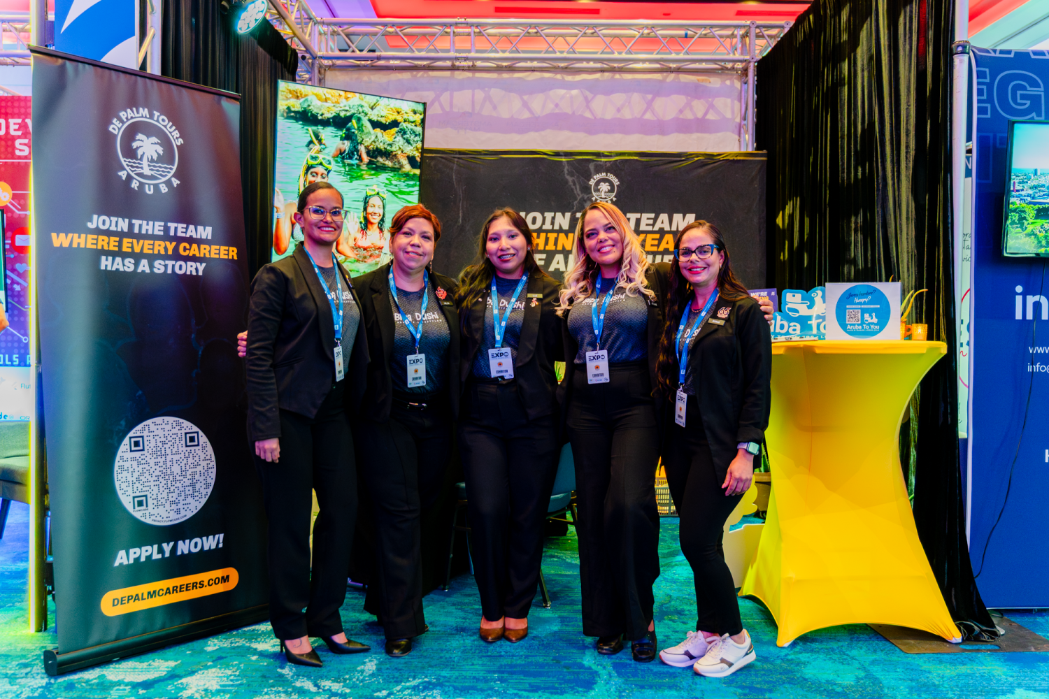 Five people in black attire stand smiling in a booth with promotional banners and materials.