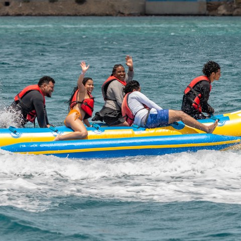 Five people in life vests riding a banana boat on the ocean, laughing and enjoying waves.
