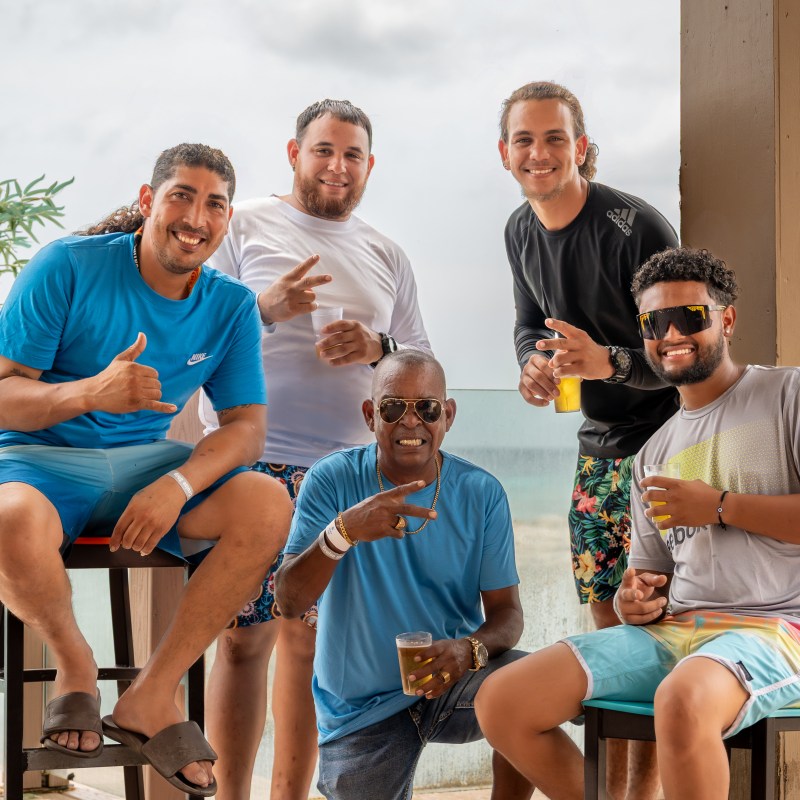 Five men smiling and posing indoors near a window with plants and drinks.
