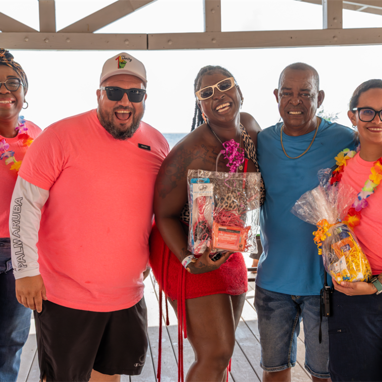 Group of people smiling in a beachside pavilion, wearing bright shirts and flower leis, holding gift bags.