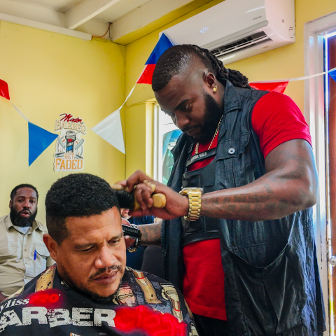Barber trimming man's hair in shop with colorful pennants hanging.