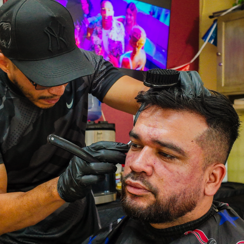 Barber wearing a cap trims a man's hair in a salon with a TV in the background.