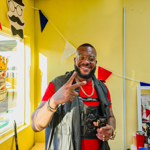 Man smiling, making peace sign in a barber shop with colorful bunting and tools.