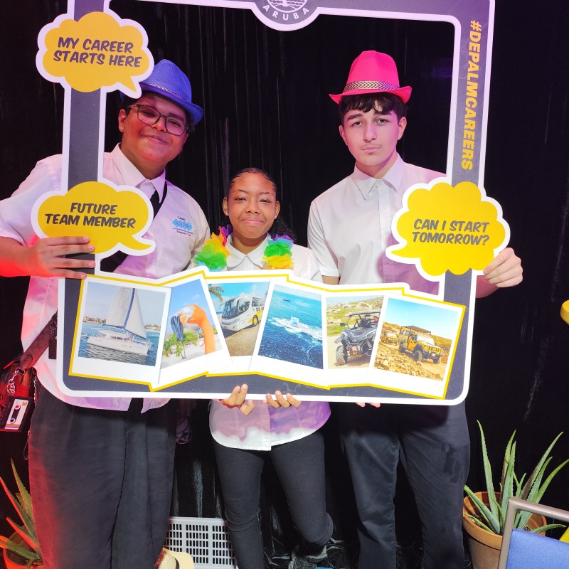 Three people in hats holding a De Palm Tours Aruba frame with speech bubbles and photos of activities.