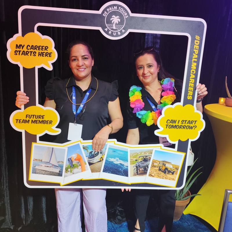 Two women holding a promotional frame for De Palm Tours Aruba at an event.