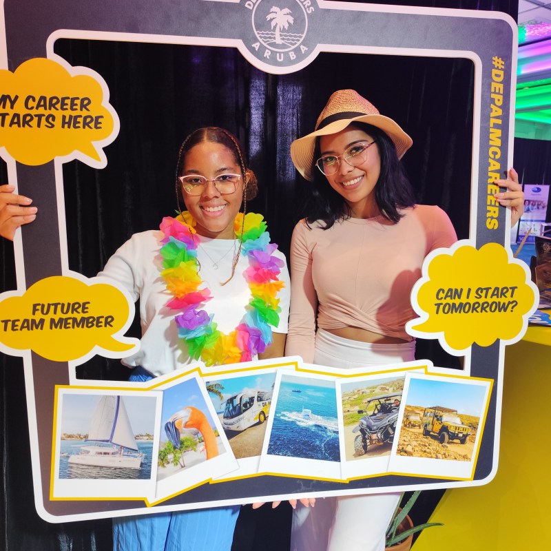 Two women posing with a De Palm Tours Aruba frame, one wearing a lei and the other a hat.