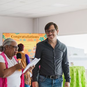 Two people standing indoors, one holding a microphone, with a 'Congratulations' sign in the background.