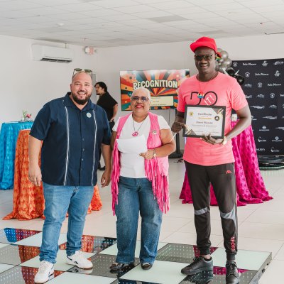 Three people posing indoors, one holding a certificate, with colorful decorations and a 'Recognition' sign behind them.