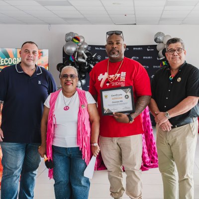Four people at a ceremony, with one holding a certificate, in front of colorful tablecloths and decorations.