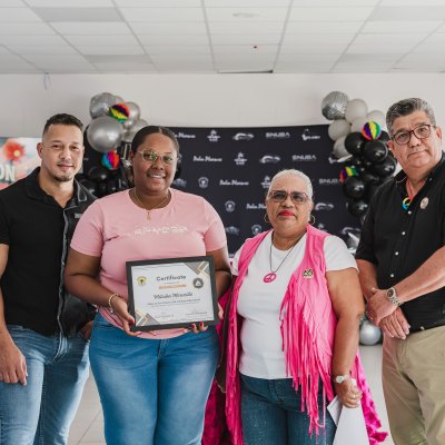 Four people standing together, one holding a certificate, with a 'Recognition' sign and balloons in the background.