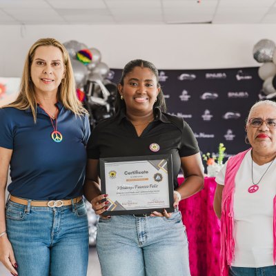 Three women standing; one holds a certificate, with balloons and a decorative background.