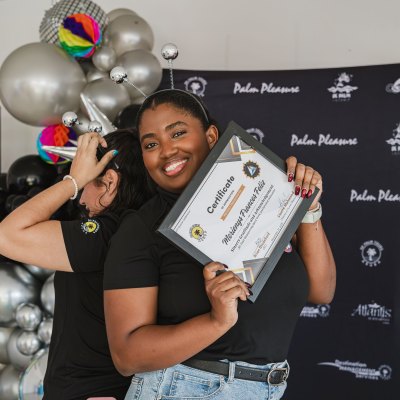 Smiling person holding a certificate with balloons and a promotional backdrop in the background.