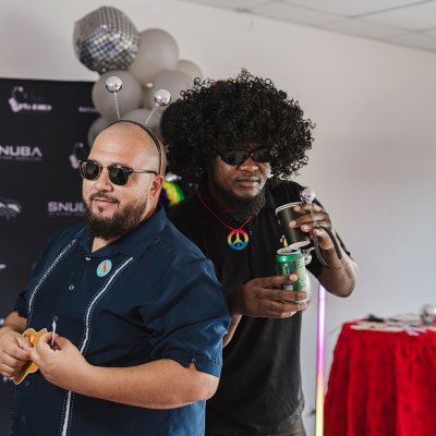 Two men in costumes and sunglasses, one holding a can, in front of a black backdrop and party decorations.