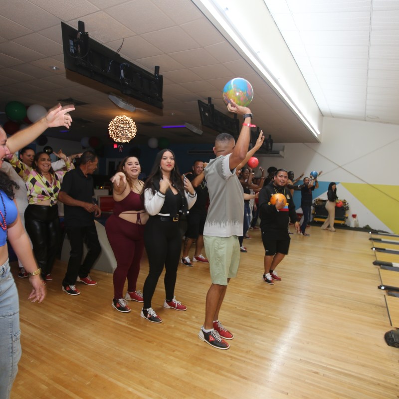Group of people dancing on a bowling alley lane, holding bowling balls.