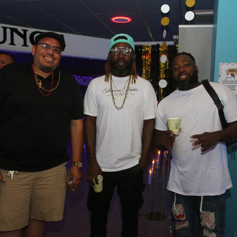 Three men smiling indoors, wearing casual clothes and holding drinks.