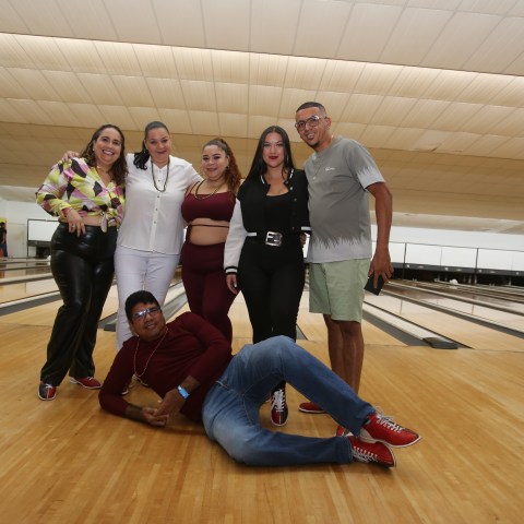 Six people posing at a bowling alley, with one person lying on the floor in front.