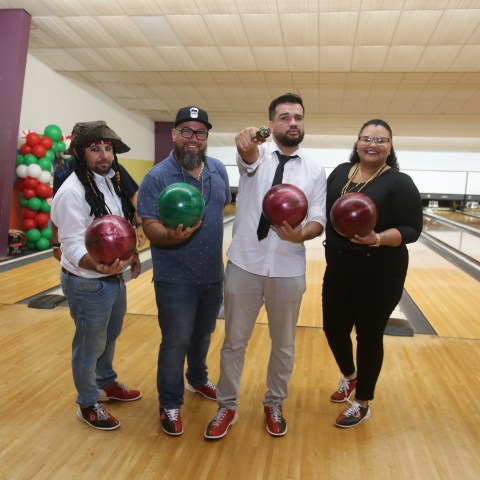 Four people holding bowling balls in a bowling alley, with colorful balloons in the background.