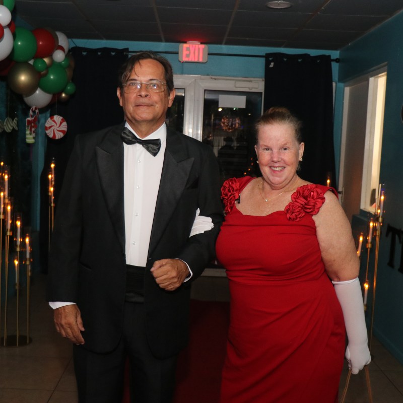 Couple in formal attire, man in tuxedo, woman in red gown, standing in decorated hallway.