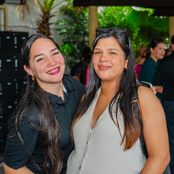 Two women smiling and posing together outdoors with people in the background.