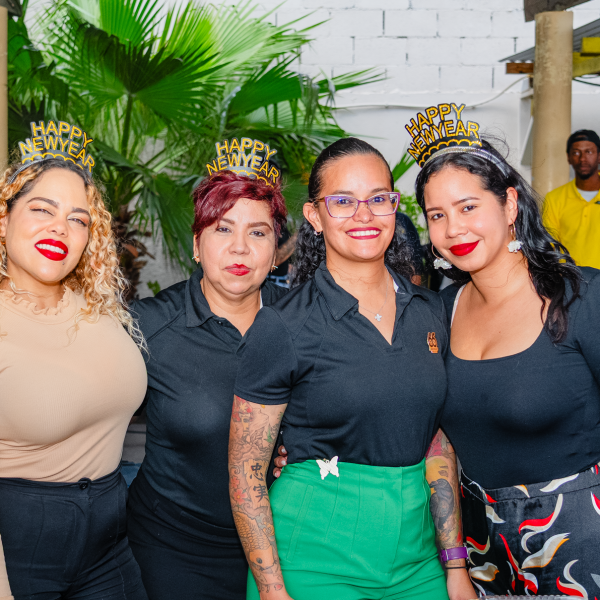 Four women wearing 'Happy New Year' headbands, standing together, smiling, with a palm plant in the background.