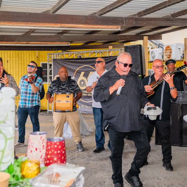 A band performs at an outdoor event under a metal roof, surrounded by tables and decorations.