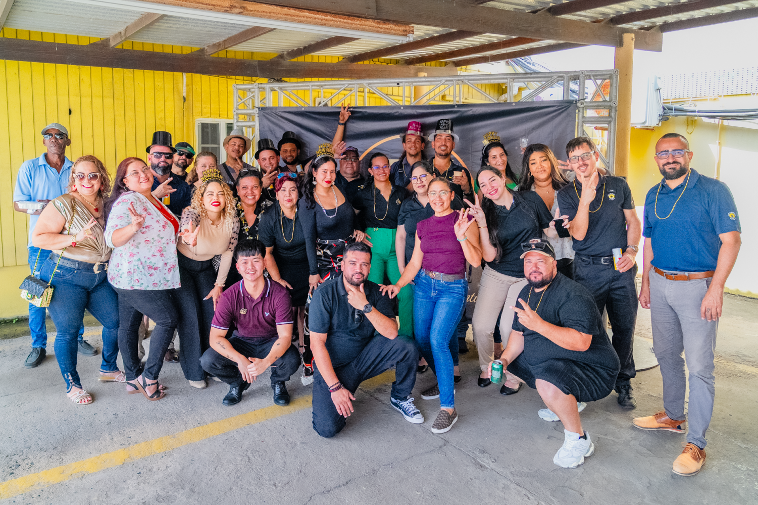 Group of people posing and smiling, wearing casual attire, in front of a yellow wall and a metal frame backdrop.