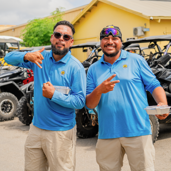 Two smiling men in blue shirts make peace signs near parked ATVs and a yellow building.