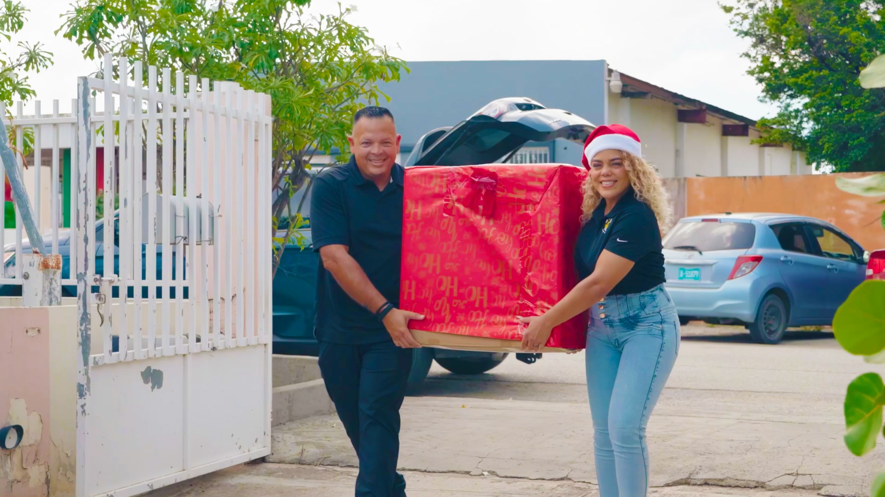 Two people carrying a large red gift box outside, one wearing a Santa hat.