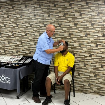 Man in blue uses device on seated man in yellow, eyewear display table nearby.