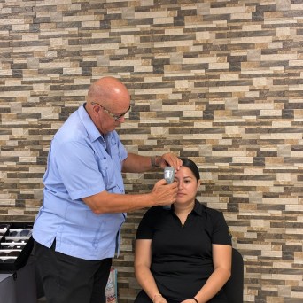 Man in blue shirt using a device on a woman's forehead in a room with tiled walls.