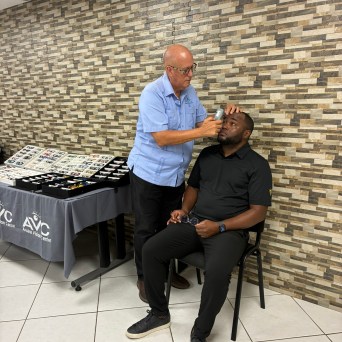 Person examining another's eye with a tool in front of a textured wall and display table.