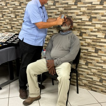 Man sitting receives eye exam from another man using a handheld device in front of a brick wall.