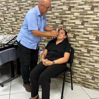 Man examines woman's eye with a device in a tiled room, eyeglass display nearby.