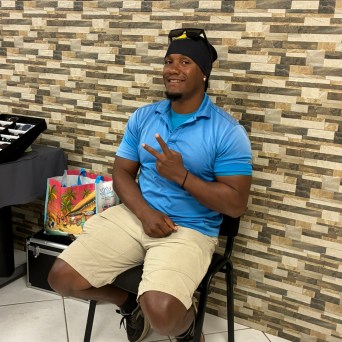 Man in blue shirt poses with peace sign, sitting on black chair against a textured wall.