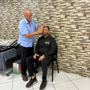 Man checking another's eyes with device against a brick-pattern wall.