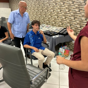 Three people in a room with a display table; one seated, two standing.